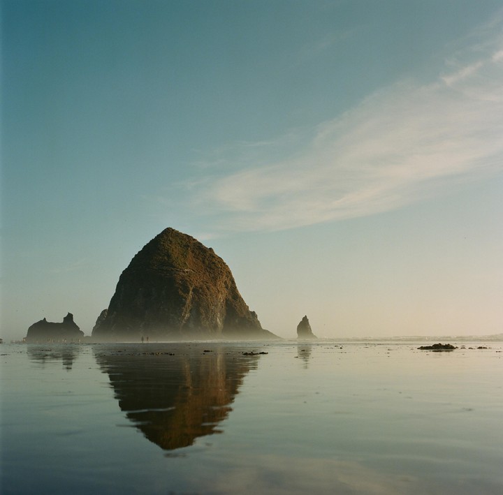 A large rock formation is reflected in the wet sand of a beach under a hazy sky.