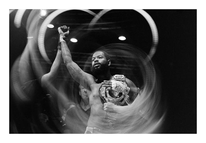 A man with a beard and tattoos raises his arm in victory while holding a championship belt.