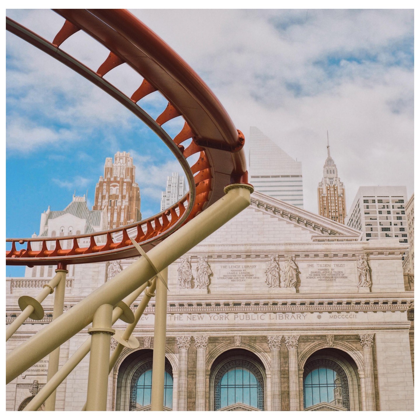 A roller coaster track curves over the New York Public Library building with other skyscrapers in the background.