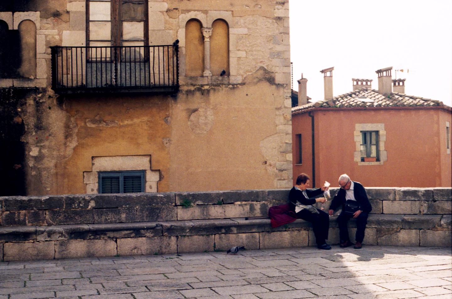 A couple sits on a stone bench in front of old buildings, with a pigeon on the ground nearby.
