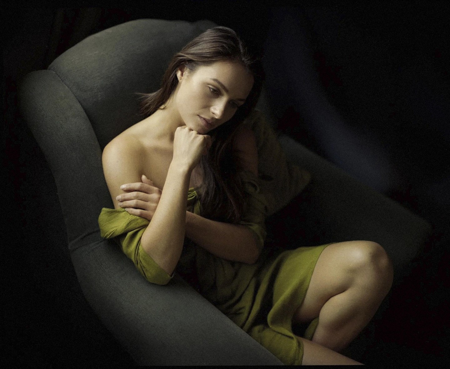 A woman in a green dress sits in a chair with her arms crossed and her hand on her chin.