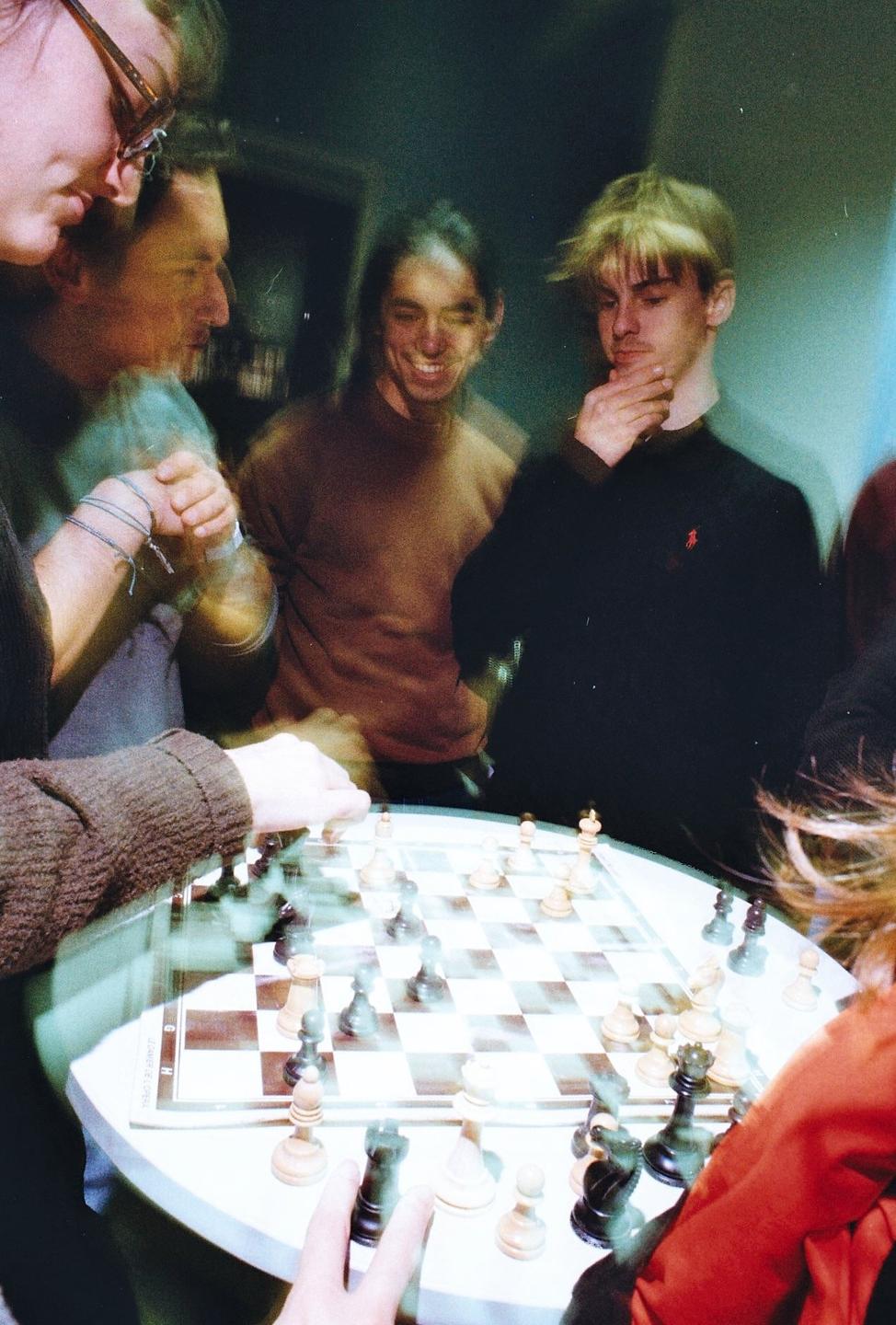 A group of men are gathered around a table playing chess in a blurry, dreamlike image.