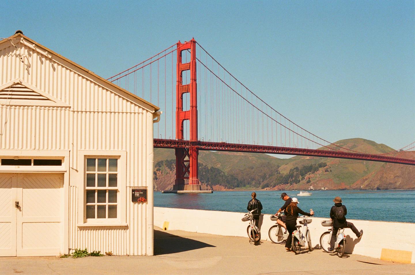A group of people on bicycles are gathered near a white building with the Golden Gate Bridge in the background.