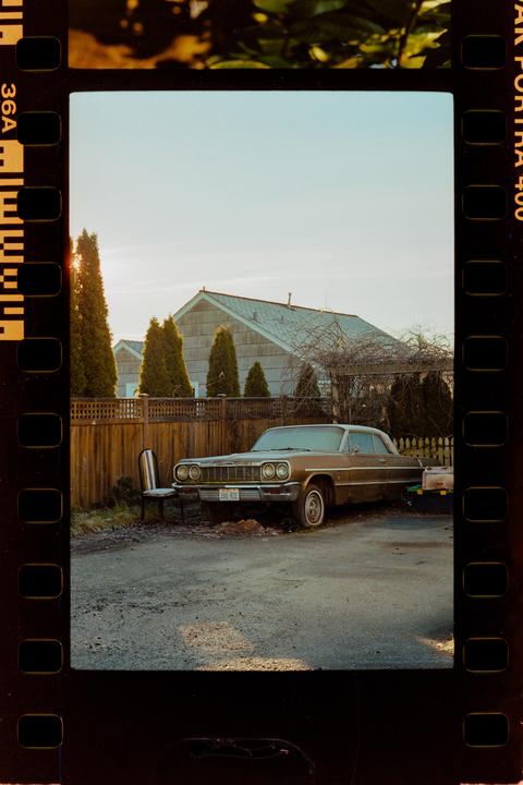 A vintage car is parked on a gravel driveway next to a wooden fence and a house.