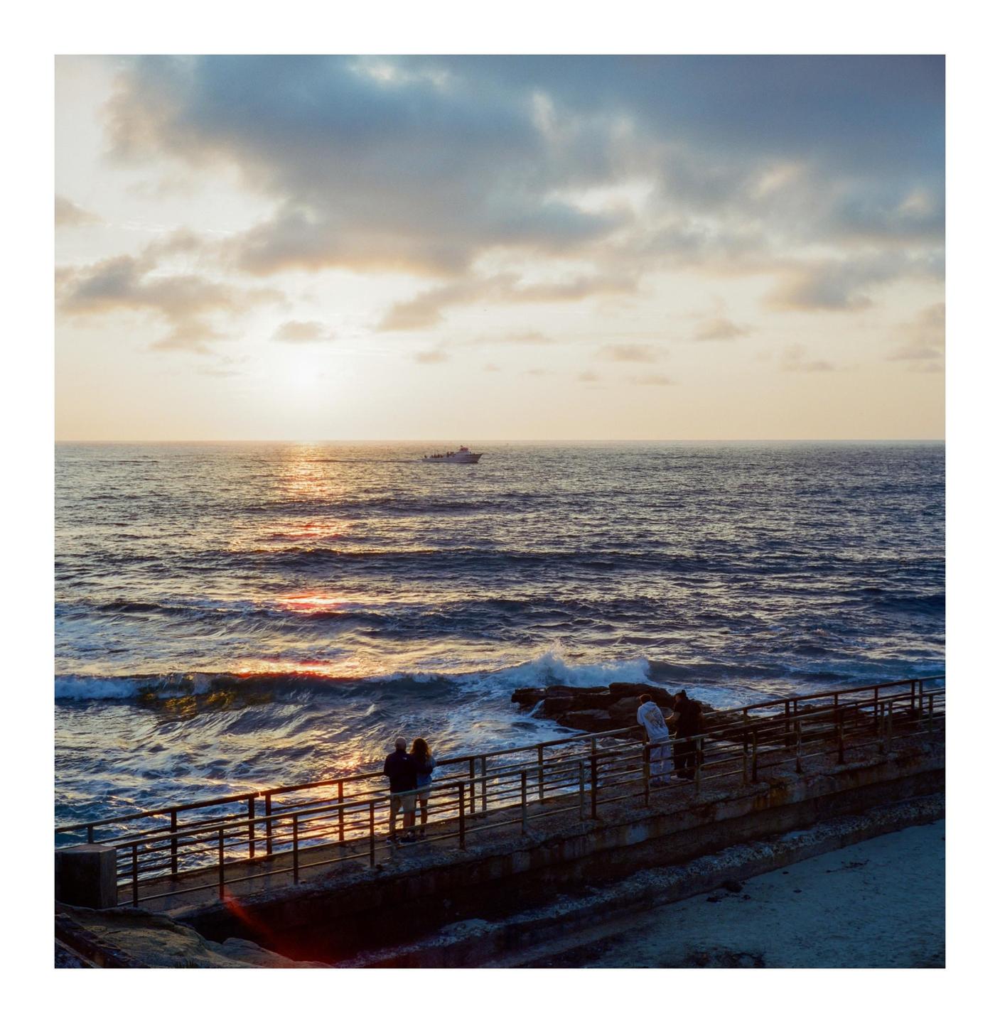 A couple and a man stand on a concrete pier with a metal railing, looking out at the ocean with a boat in the distance under a cloudy sunset sky.