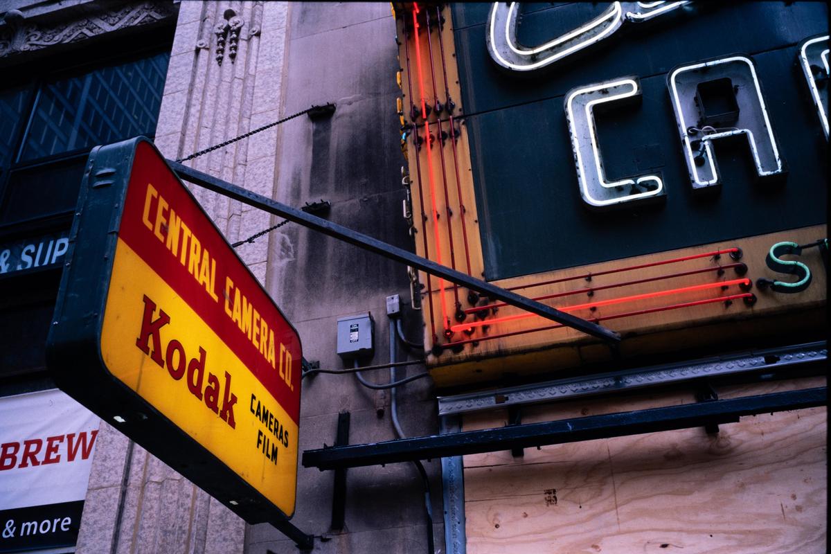 A yellow and red sign for Central Camera Co. and Kodak hangs from a building next to a neon sign.