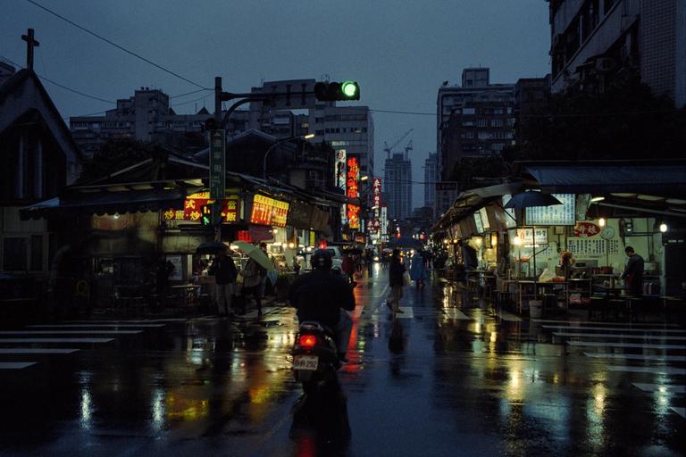 A street in Taipei is wet from rain, with neon signs illuminating the night and a person on a scooter driving away from the viewer.