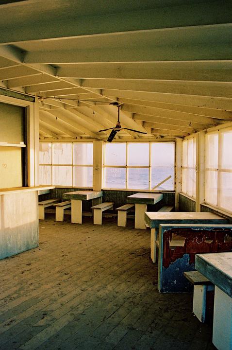 An empty, weathered room with wooden tables and benches overlooks the ocean through large windows.