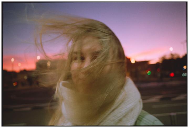 A woman with her hair blowing across her face is visible against a colorful sunset sky.