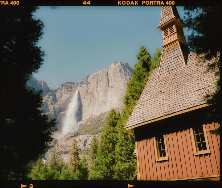 A wooden chapel with a tall steeple stands among trees, with a large waterfall and mountain in the background.