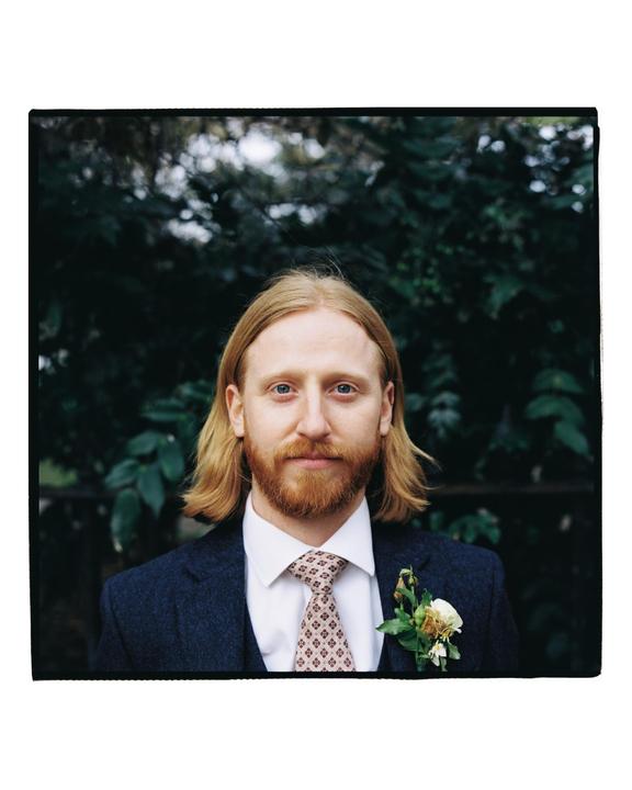 A man with long blond hair and a red beard wears a suit and tie with a boutonniere.