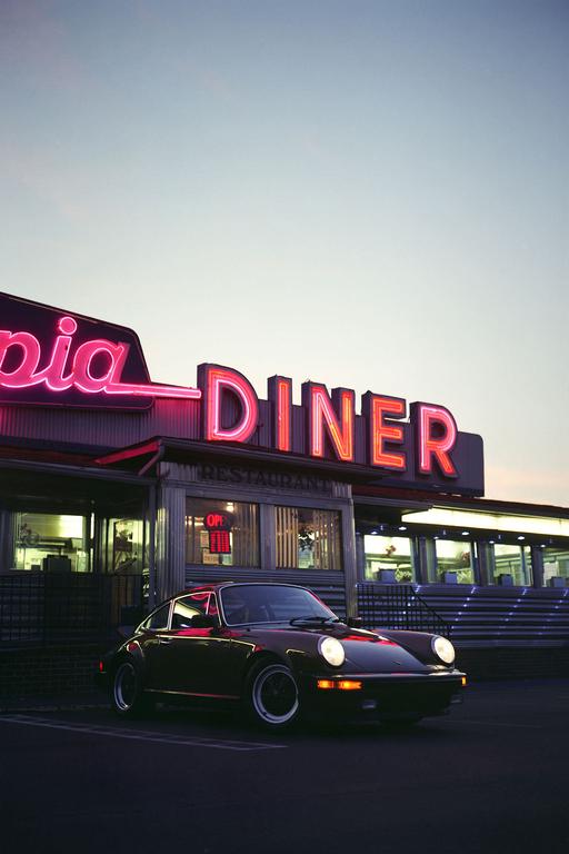 A black car with its headlights on is parked in front of a diner with neon signs at dusk.