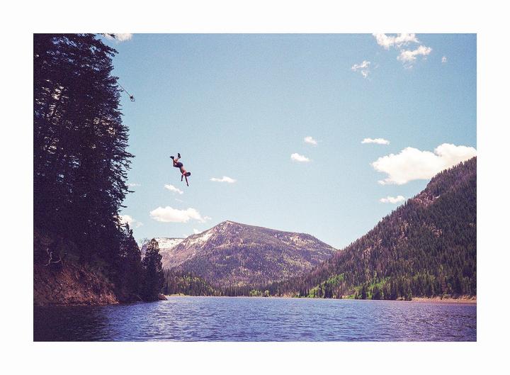 A man is captured mid-air as he dives into a lake surrounded by mountains and trees.