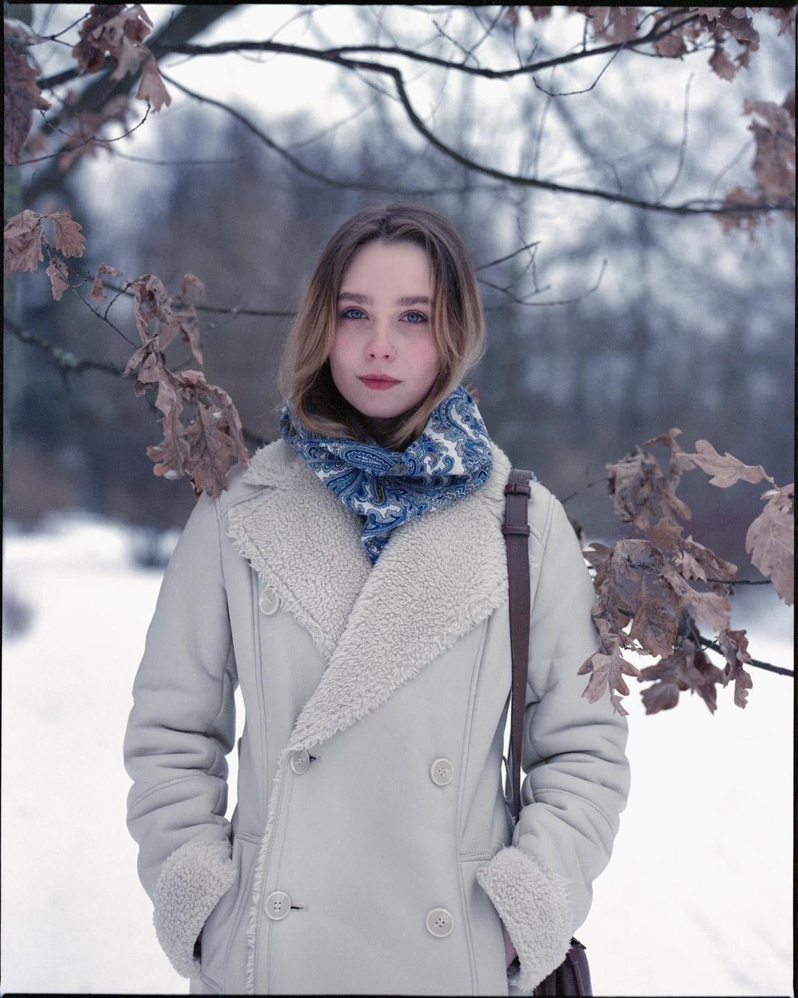 A woman wearing a coat and scarf stands in a snowy landscape with bare tree branches and dried leaves.