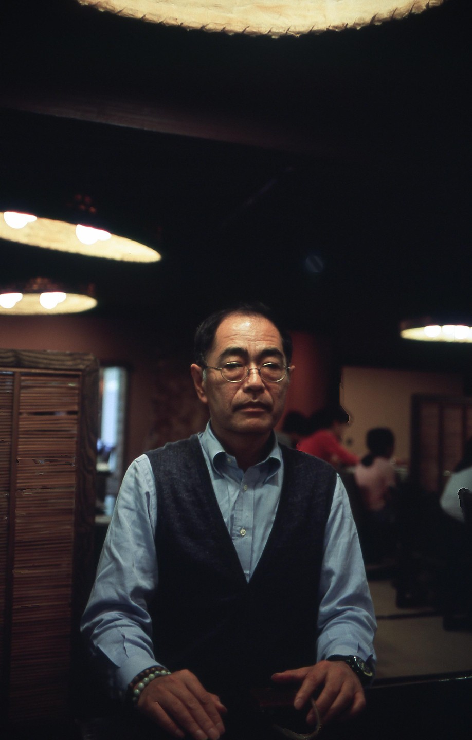 A man wearing glasses and a vest sits in a dimly lit room with circular lights on the ceiling.