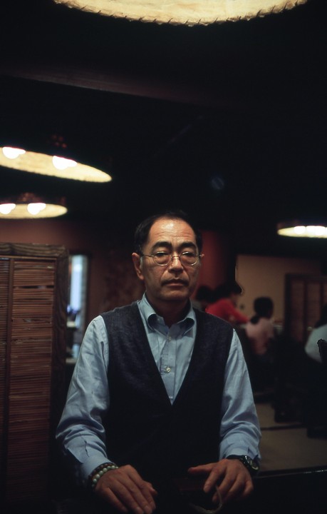 A man wearing glasses and a vest sits in a dimly lit room with circular lights on the ceiling.