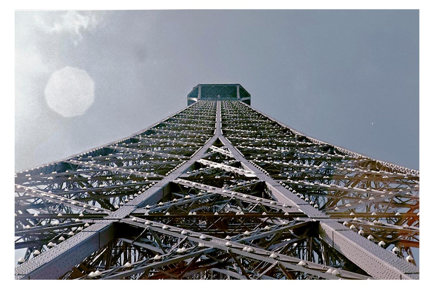 The Eiffel Tower is viewed from directly below, looking up at its intricate metal structure against a bright sky.