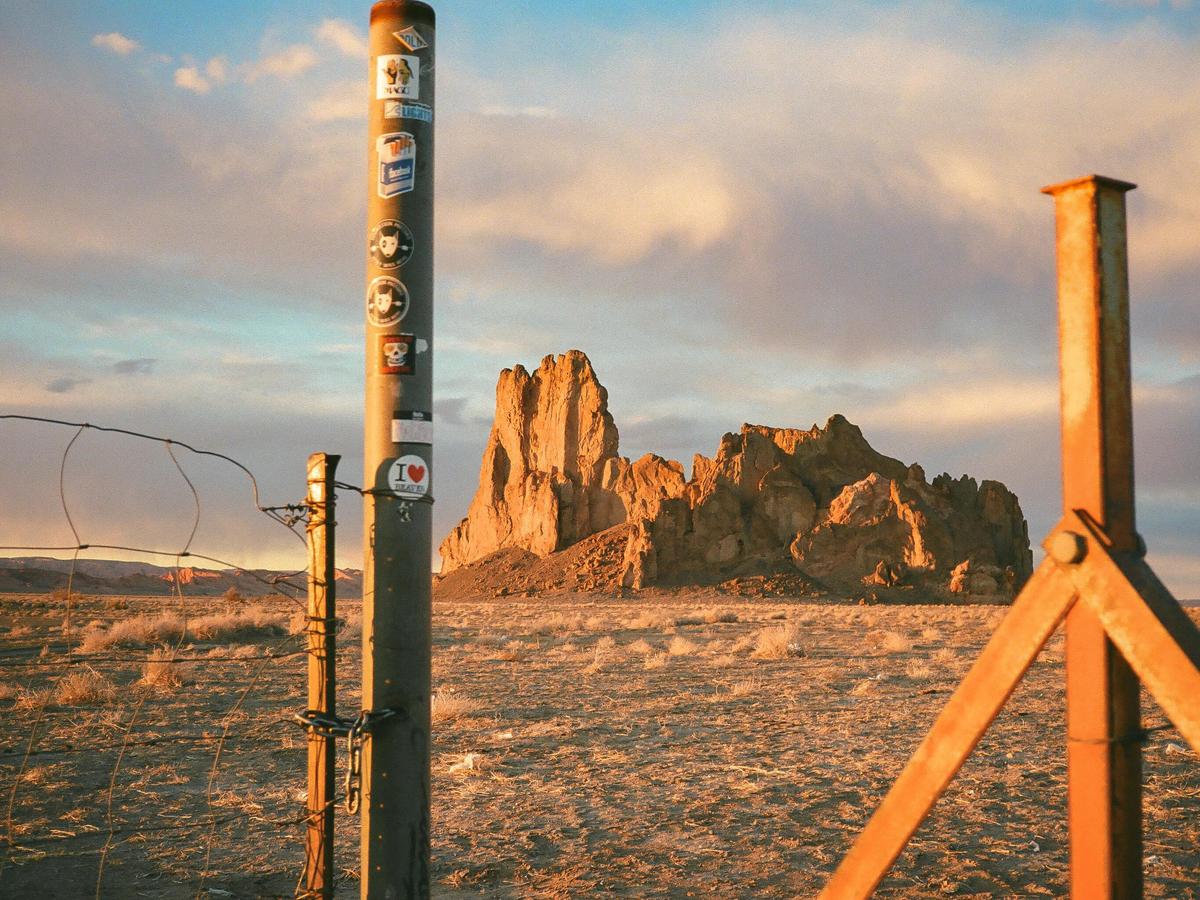 A fence post with stickers stands in the foreground of a desert landscape with a large rock formation under a cloudy sky.