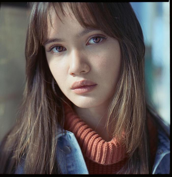 A woman with long hair and bangs wears a turtleneck sweater and denim jacket.