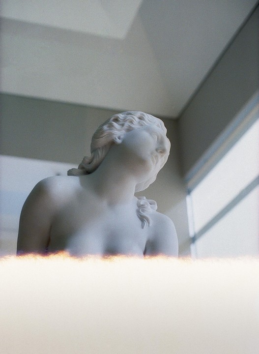 A white marble statue of a woman is shown from the chest up, looking towards a window with light streaming in.