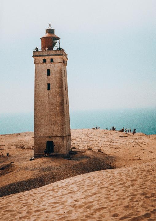 A tall, weathered lighthouse stands on a sandy dune with the ocean visible in the background and people gathered around.