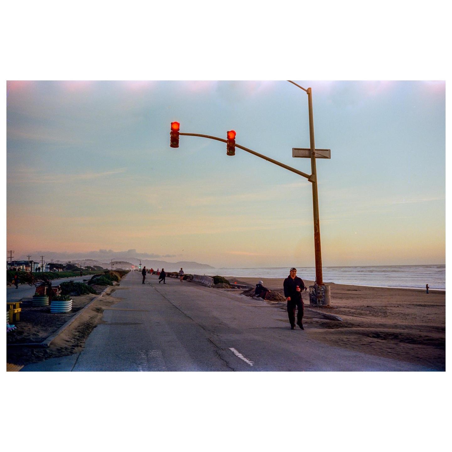 A man walks along a coastal road with a beach and ocean to his right, and a traffic light overhead.