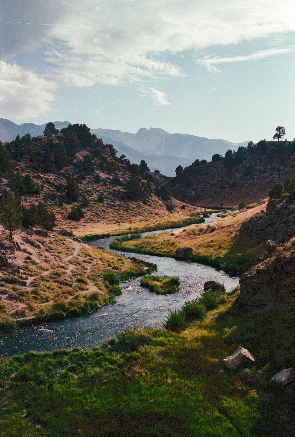A winding river flows through a valley with hills and mountains in the background under a cloudy sky.