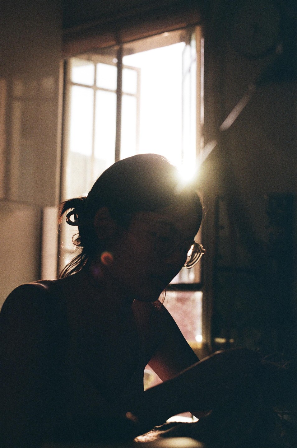 A woman with her hair tied back and wearing glasses is silhouetted against a bright window.