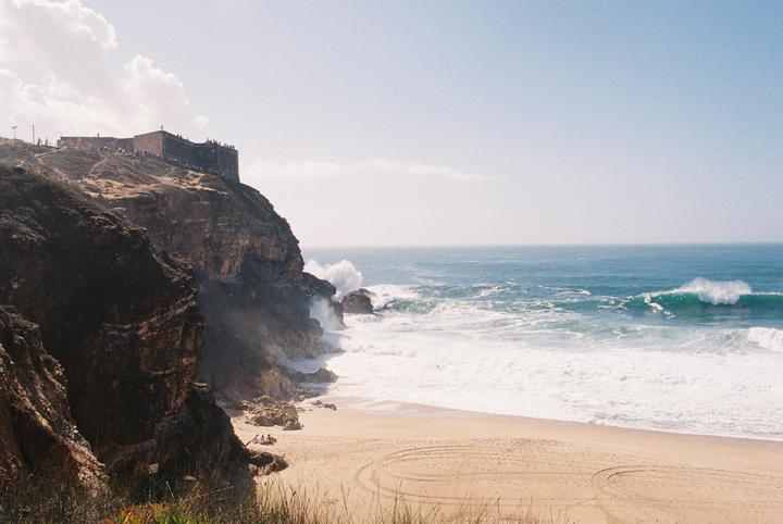 A large cliff with a building on top overlooks a sandy beach with crashing waves.