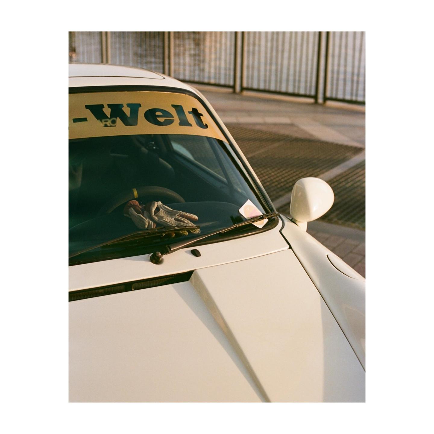 A white car with a yellow banner on the windshield and a glove on the dashboard is parked outdoors.