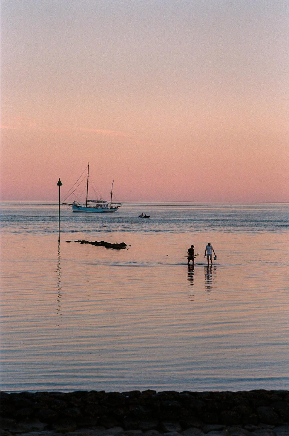 Two men wade in shallow water with a sailboat and a small motorboat in the background under a pink sky.