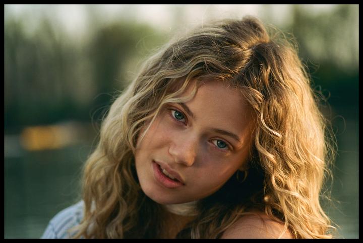 A young woman with curly blonde hair and blue eyes looks directly at the viewer.