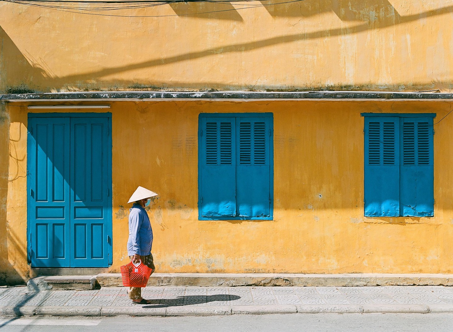 A woman wearing a conical hat and a face mask walks past a yellow building with blue doors and windows.