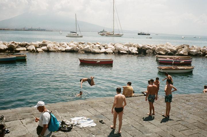 A boy dives into the water from a stone pier where other boys are gathered, with boats and a mountain in the background.