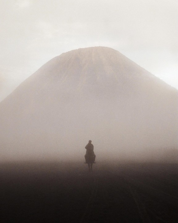 A man on horseback rides through a foggy landscape with a mountain in the background.