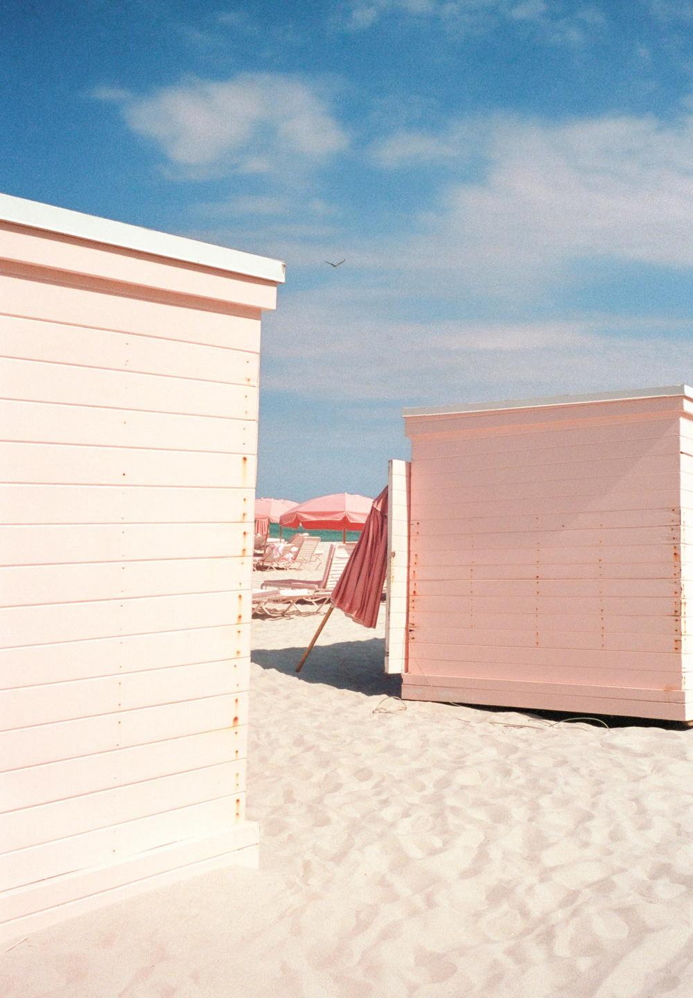 Two pink beach huts stand on a sandy beach with pink umbrellas and lounge chairs in the background under a blue sky.