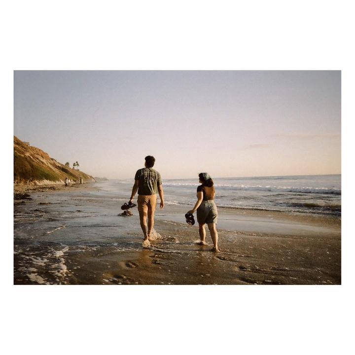 A couple walks barefoot along the water's edge on a beach with a cliff on the left.