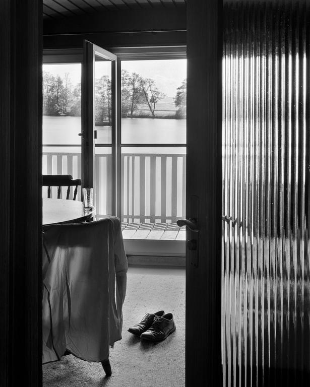A pair of shoes sits on the floor next to a chair with a shirt draped over it, with a view of a lake and trees through an open door.