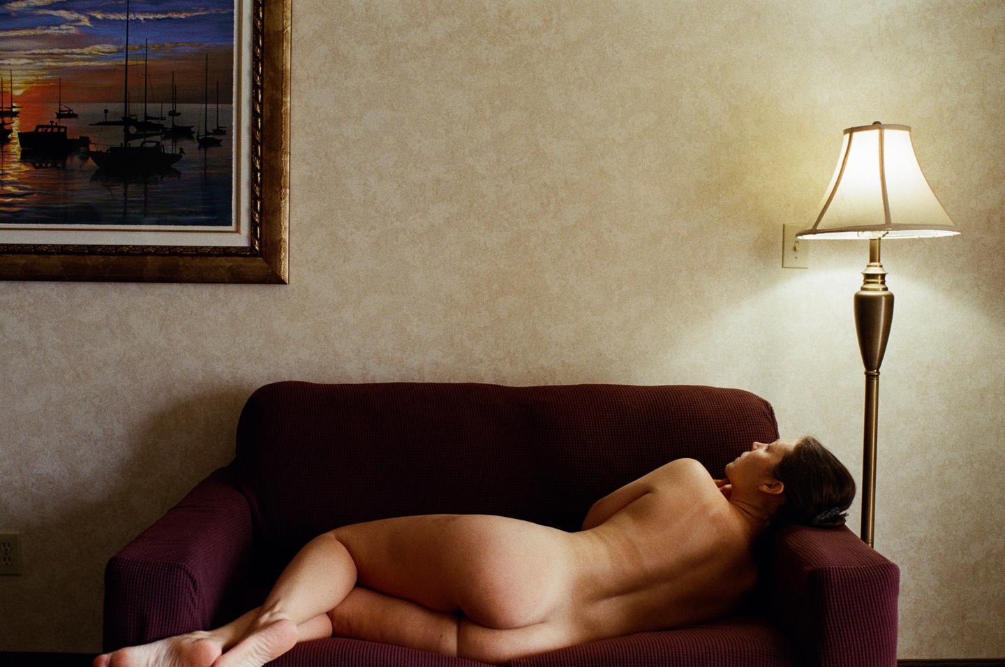 A woman lies nude on a sofa next to a floor lamp, with a painting of boats at sunset on the wall.