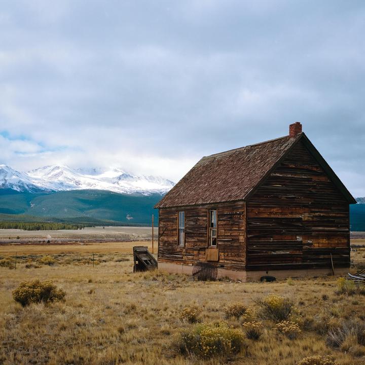 An old wooden house stands in a field with snow-capped mountains in the background under a cloudy sky.
