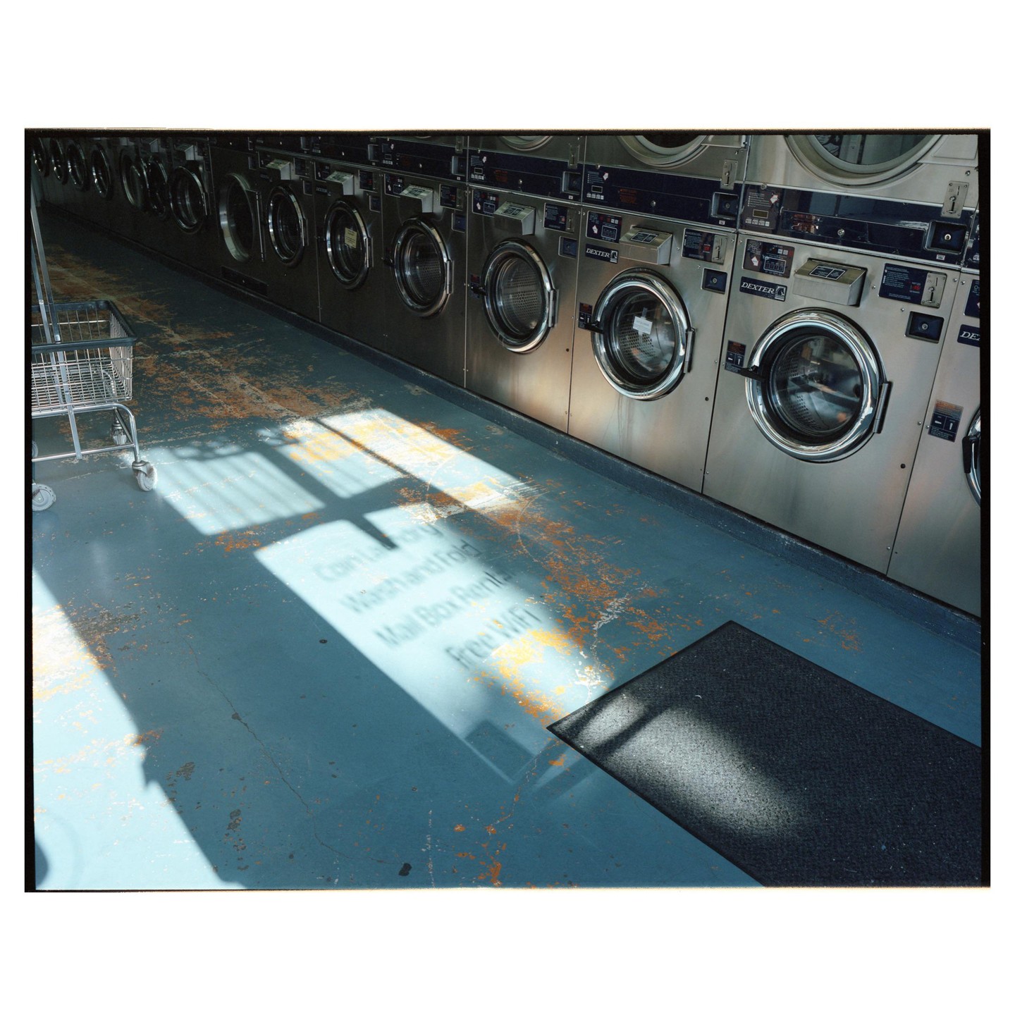 Sunlight streams across the floor of a laundromat with a row of washing machines and a laundry cart.