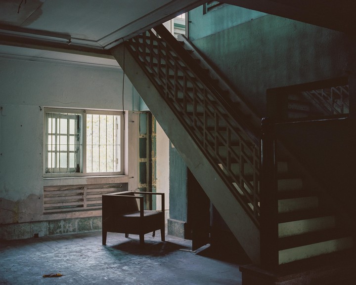A wooden chair sits in a derelict room with a barred window and a staircase leading upwards.