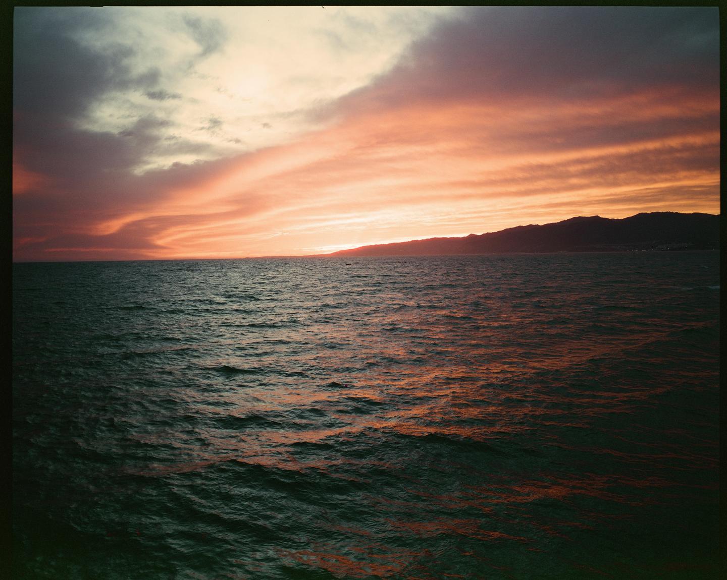 A vibrant sunset casts orange and pink hues across the sky and reflects on the ocean, with a dark mountain range visible in the distance.