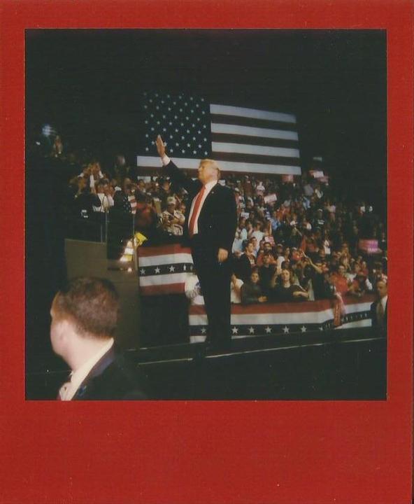 A man in a suit waves to a crowd in front of a large American flag.