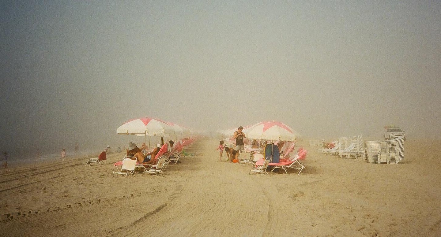 People relax under umbrellas on a hazy beach with a lifeguard truck in the distance.