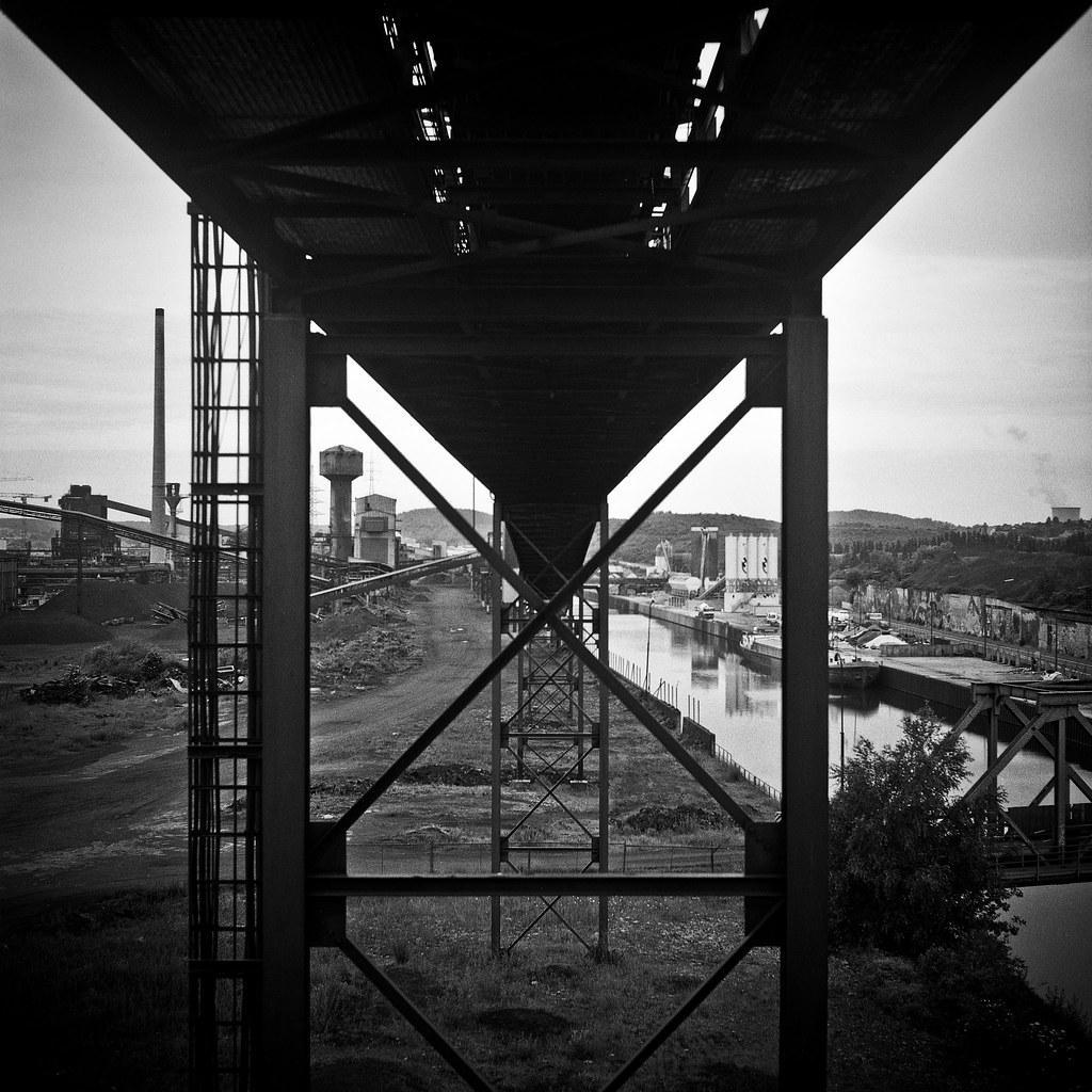 A black and white photograph shows an industrial landscape with a canal, factories, and a large metal structure in the foreground.