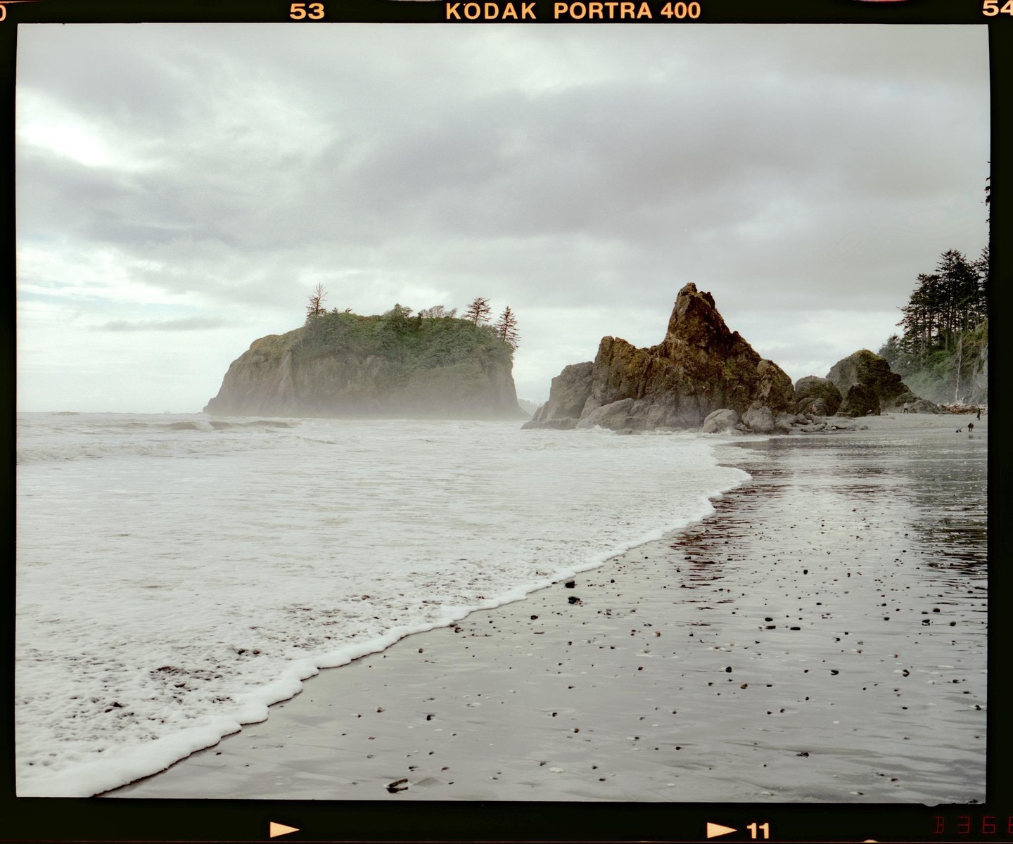 Waves crash on a sandy beach with large rock formations and an island covered in trees under a cloudy sky.