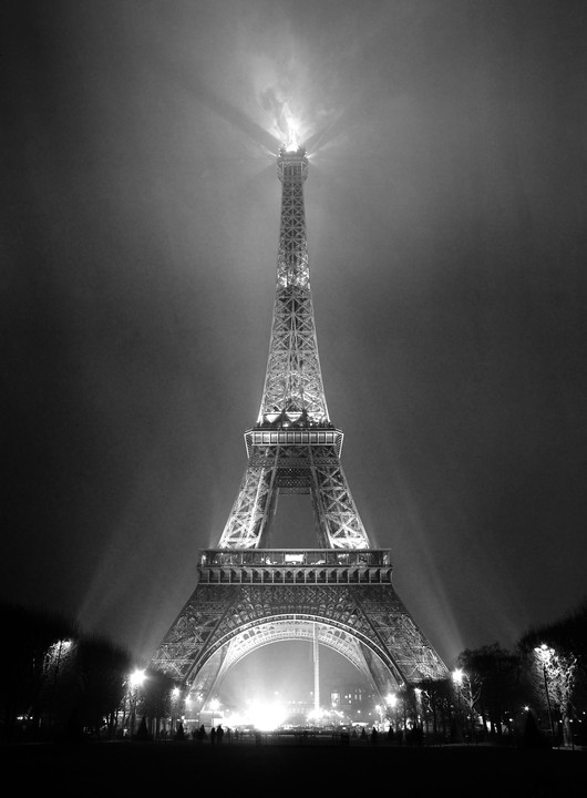 The Eiffel Tower is illuminated at night, with light beams extending into the hazy sky.