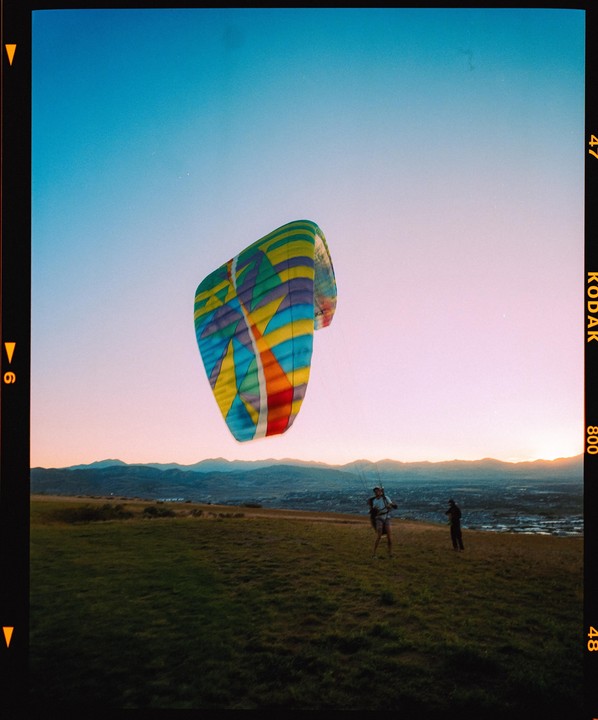 Two men are on a grassy hill with a colorful paraglider canopy above them at dusk, with mountains in the background.