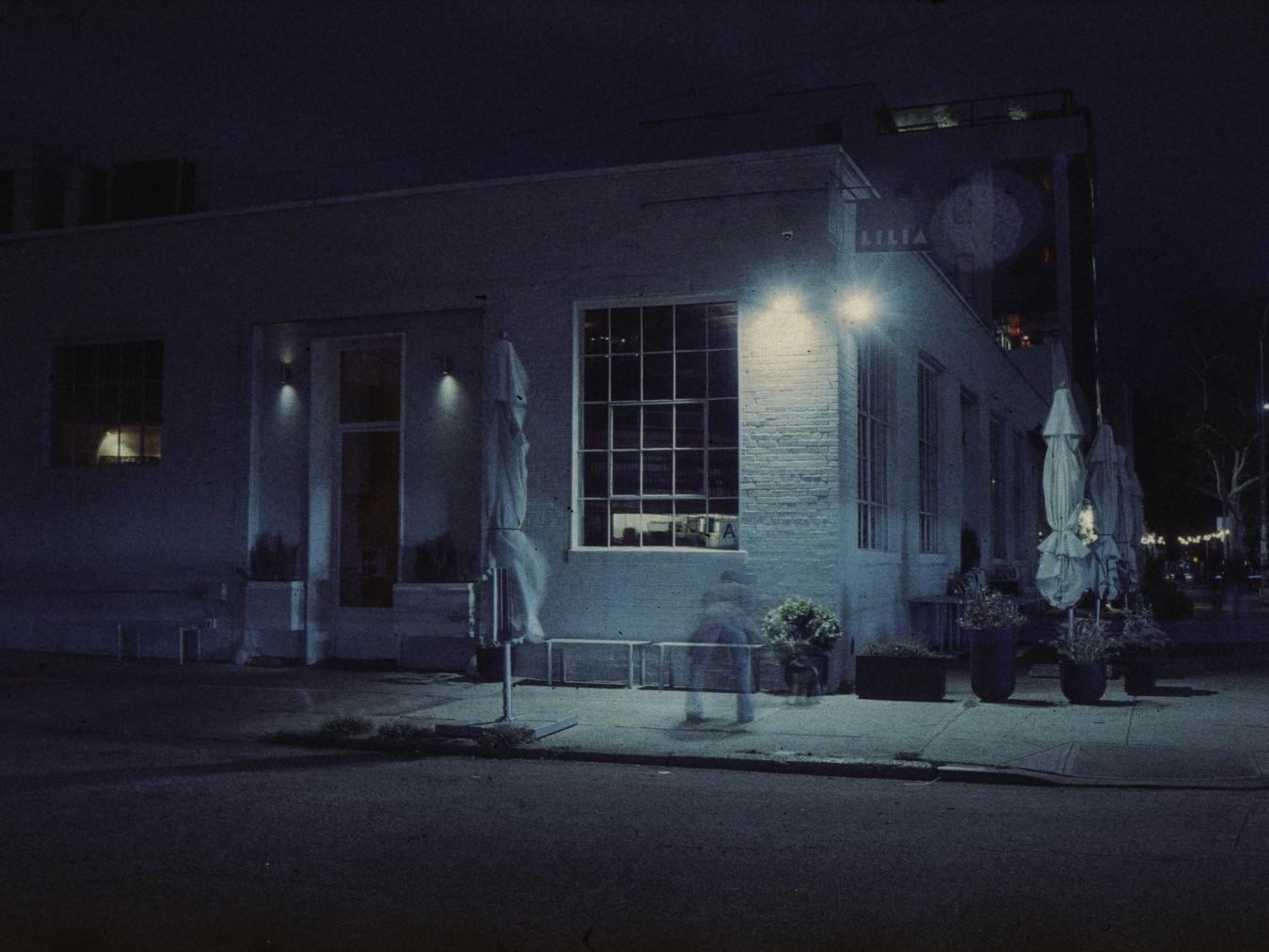 A blurred figure walks past a building with large windows and covered outdoor umbrellas at night.